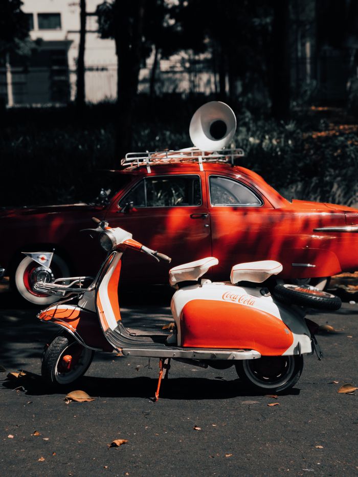 A nostalgic scene featuring a red vintage scooter and car in an outdoor setting with a moody tone.